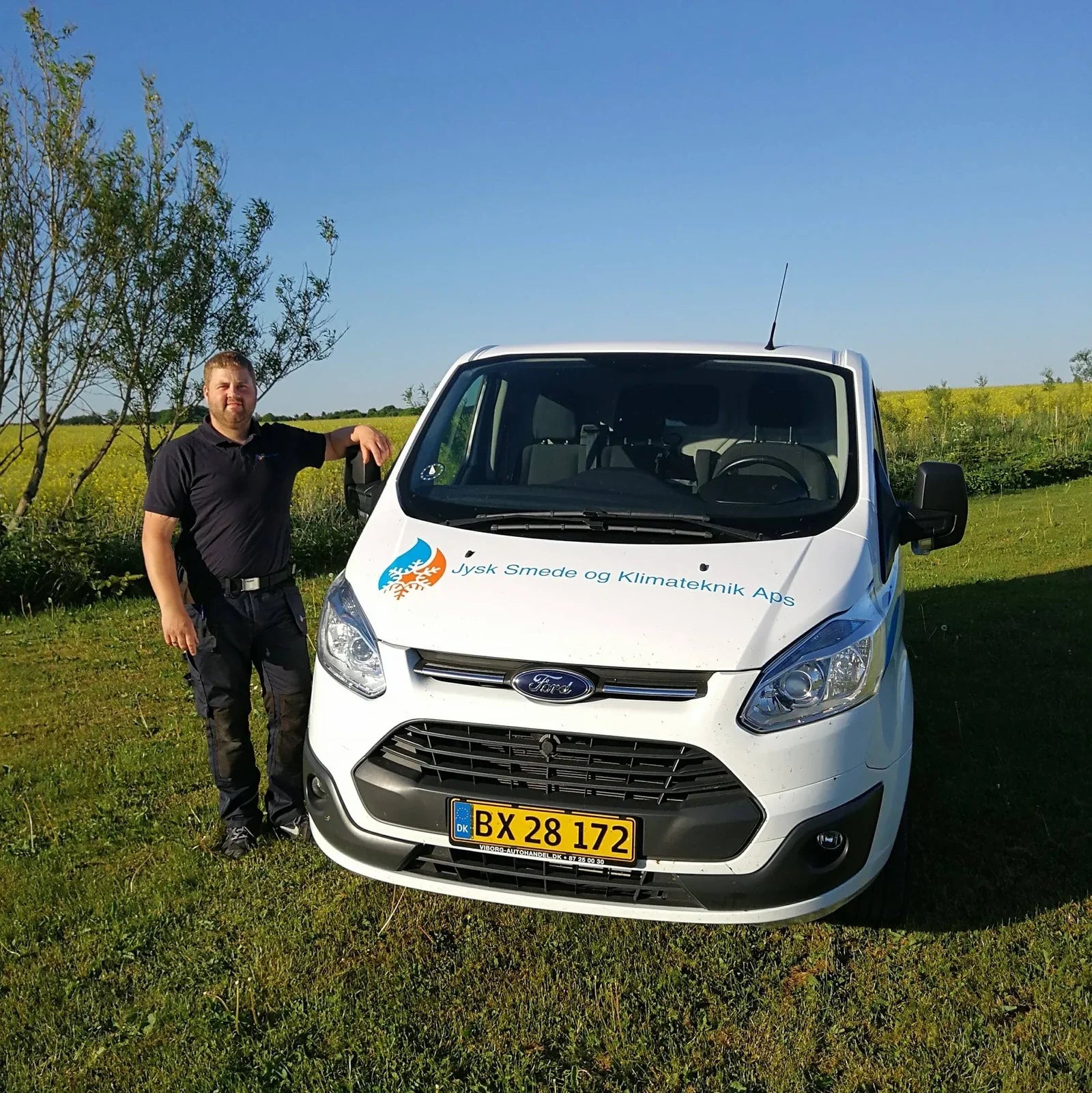 Hvid firmabil fra Jysk Smede og Klimateknik ApS med logo, parkeret på græs ved mand i arbejdstøj.